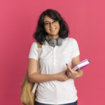 young smiling pretty caucasian schoolgirl wearing glasses headphones and back bag holds book isolated on pink background with copy space